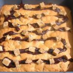 A close-up of a choc chip fudge madeira traybake drizzled with melted chocolate and topped with pieces of fudge.