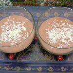 Two glass bowls of Greek yoghurt chocolate pudding garnished with white chocolate flakes. Sitting on a purple Mexican style tray.