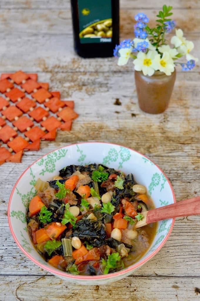 A bowl of ribollita: a hearty vegan Tuscan soup - with spoon. Flowers, a red mat and partial view of a bottle of olive oil completes the picture.