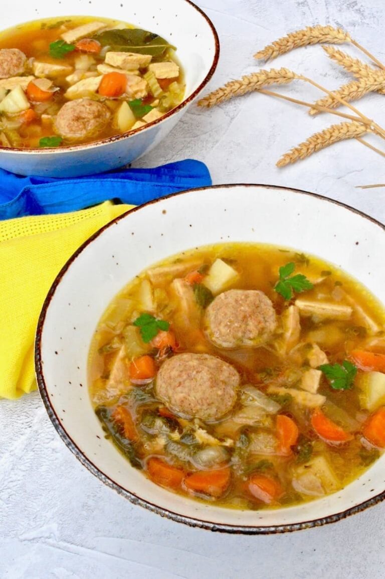 A bowl of vegetable soup with two dumplings in the foreground. Blue and yellow napkins representing the Ukrainian flag, some ears of wheat and a second bowl in the background.