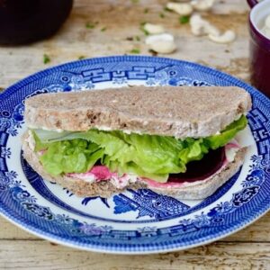 Vegan 'Cream Cheese', Spiced Pickled Beetroot & Lettuce Sandwich on a blue plate. Cashew nuts and other paraphernalia in the background.