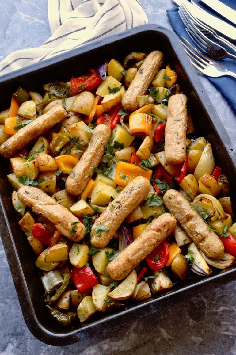 A one pan vegan sausage traybake with peppers and potatoes in roasting tin. Striped cloth, navy napkins and a pile of knifes and forks in the background.