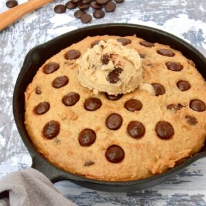 A giant vegan chocolate chip skillet cookie with a scoop of chocolate ice-cream on top. A wooden spoon amidst a load of chocolate chips completes the picture.