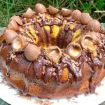 A spiced chocolate & orange Easter bundt cake drizzled with chocolate sauce and decorated with mini Easter eggs and sitting on a white plate.