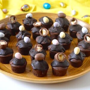 Mini spiced rum Easter chocolate cakes topped with a whole or half chocolate egg. Sitting on a bamboo tray with various mini Easter eggs in the background..