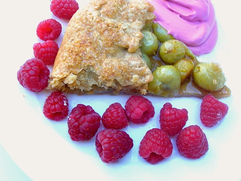 A slice of gooseberry galette on a white plate with raspberries and blueberry cream on the side.