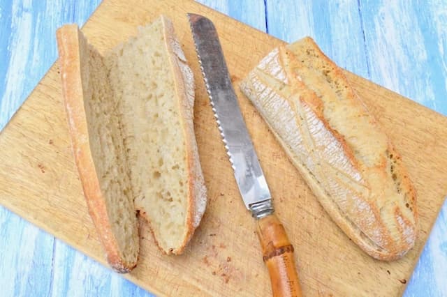 A bread board with two demi baguette halves sliced, but not completely. A bread knife in the middle.