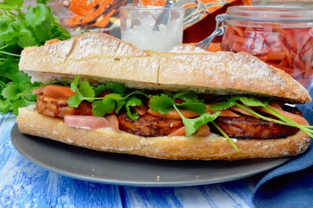 A close up of the middle section of a vegan tempeh bánh mi Vietnamese sandwich. A jar of mayonnaise and coriander (cilantro) leaves in the background.