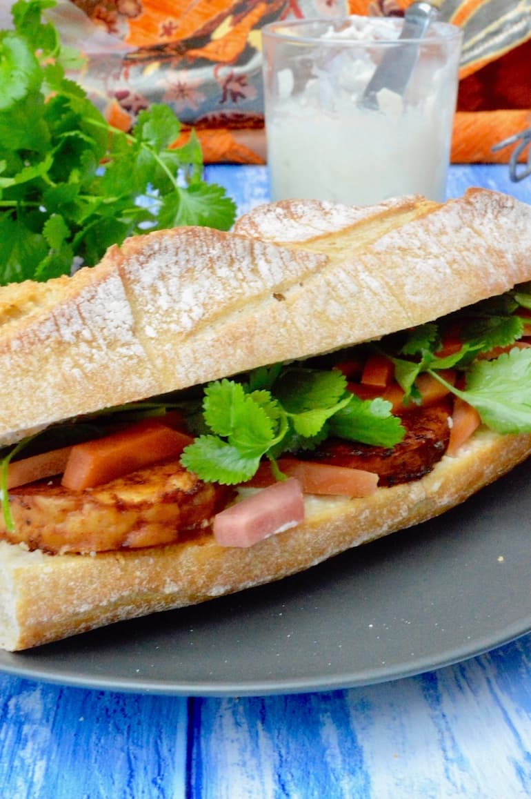 A close up of the middle section of a vegan tempeh bánh mi Vietnamese sandwich. A jar of mayonnaise and coriander (cilantro) leaves in the background.
