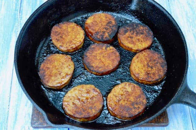 Marinated tempeh slices frying in a cast iron skillet.