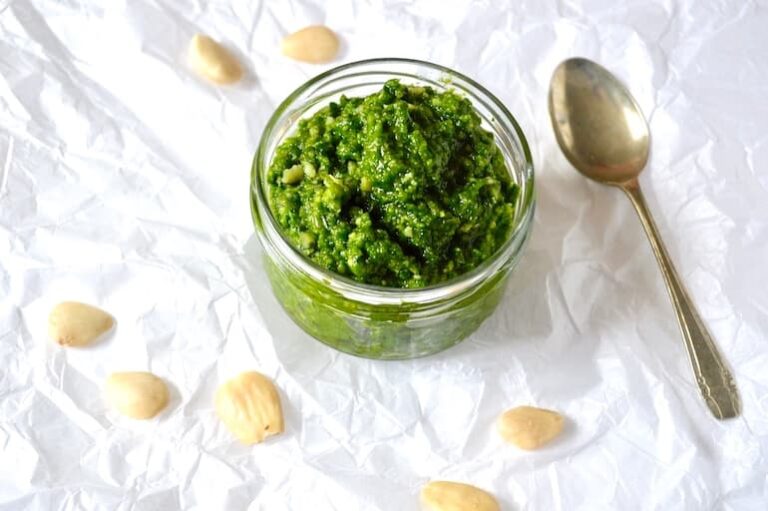 An open jar of wild garlic pesto with spoon and almonds scattered around.