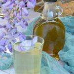 A bottle of wisteria syrup with a glass of cordial at the front, wisteria flowers on the side and green gauze in the background.