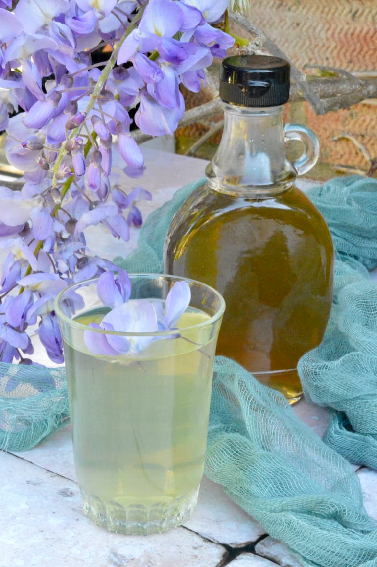 A bottle of wisteria syrup with a glass of cordial at the front, wisteria flowers on the side and green gauze in the background.