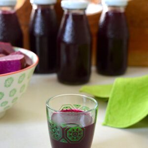 A glass of beet kvass with four bottles behind and a bowl of lacto-fermented beetroot on the side.