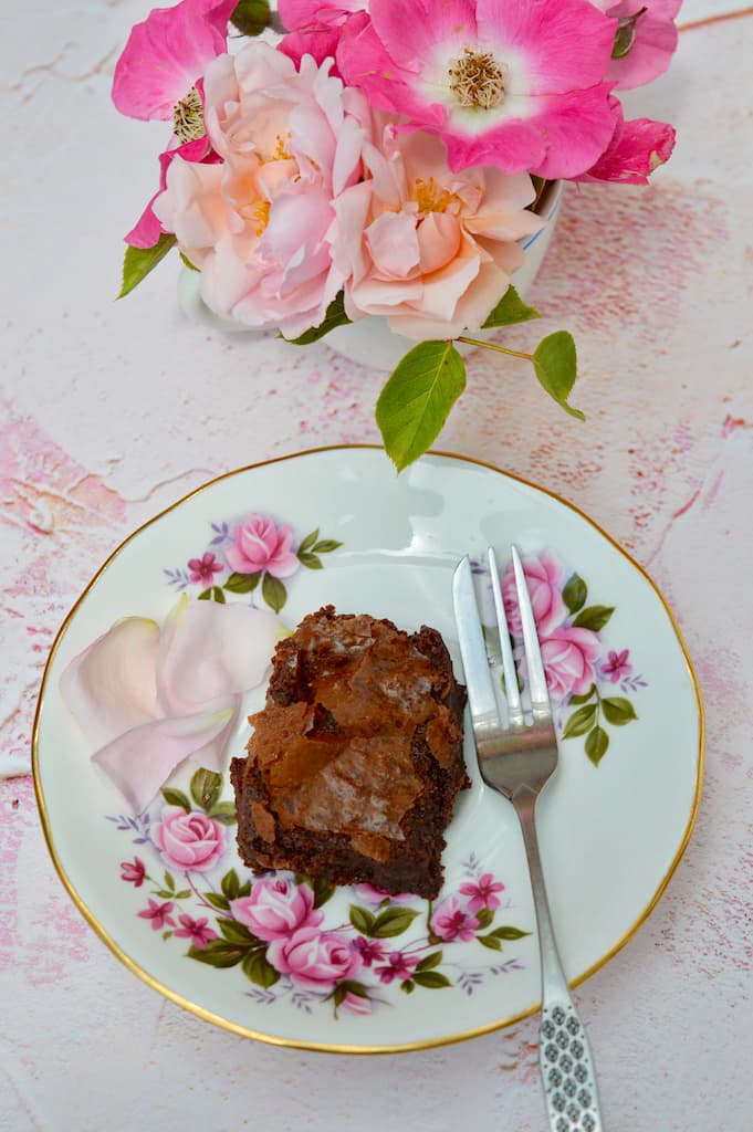 A rose cardamom brownie on a plate alongside a vase of fresh roses.