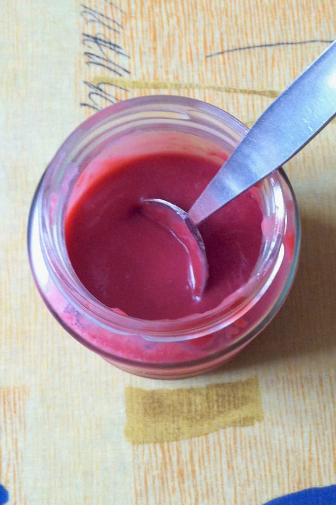 A jar of opened raspberry and rose curd with a spoon inside.