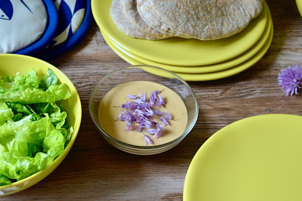 A bowl of tahini sauce with chive flowers scattered over the top. Set amongst yellow plates, bread and lettuce.