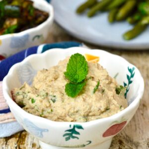 A bowl of baba ganoush with a sprig of mint on top and a plate of beans behind.