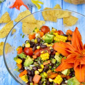 A glass bowl of black bean & corn salad decorated with a day lily and tortilla chips in the background.