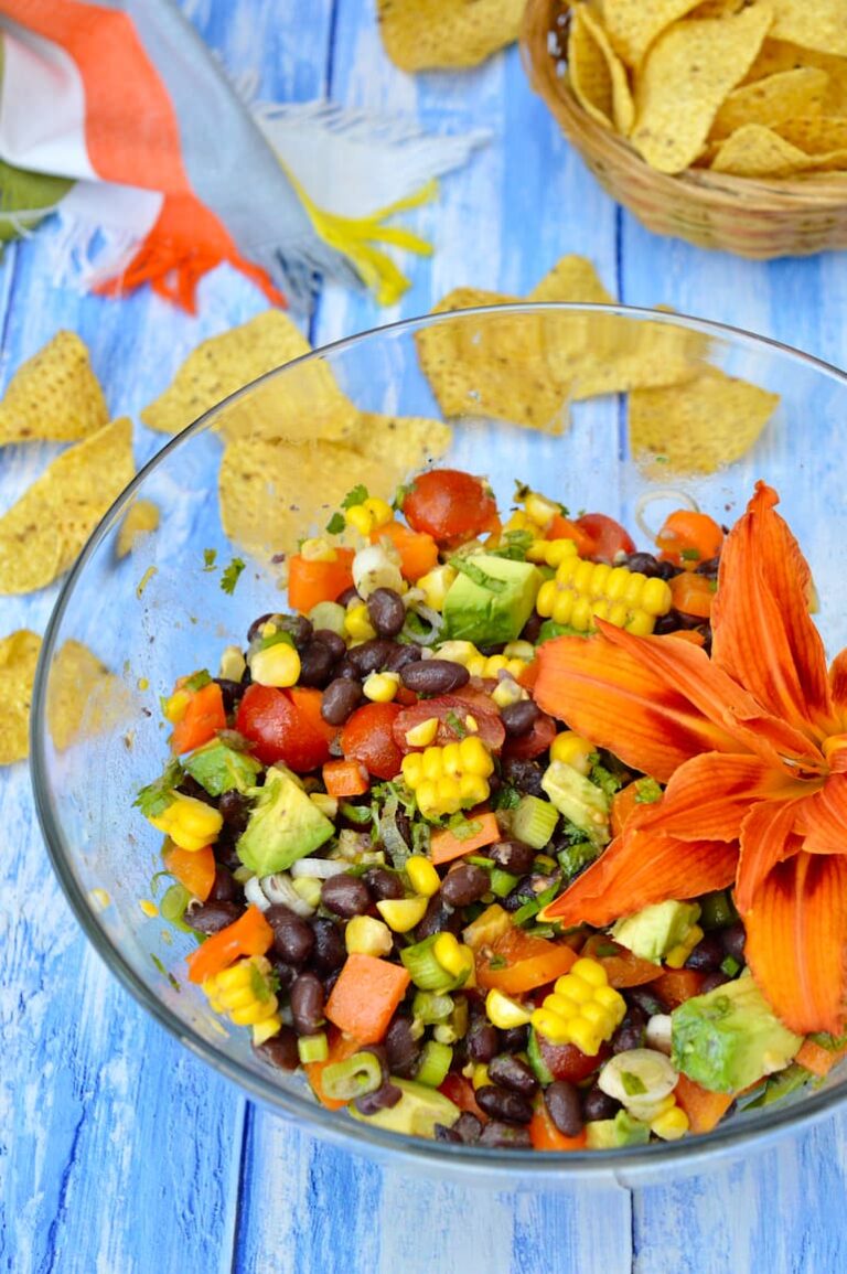 A glass bowl of black bean & corn salad decorated with a day lily and tortilla chips in the background.