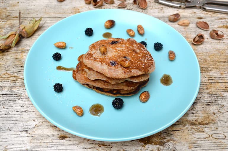 A stack of blackberry & apple spelt pancakes with brown buttered cobnuts scattered on turquoise plate.