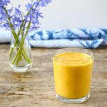 A glass of carrot smoothie with ginger and orange and a vase of bluebells behind.