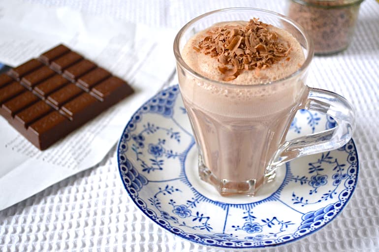 A glass of homemade indulgent chocolate milk topped with grated chocolate. A partial bar of unwrapped chocolate on the side.