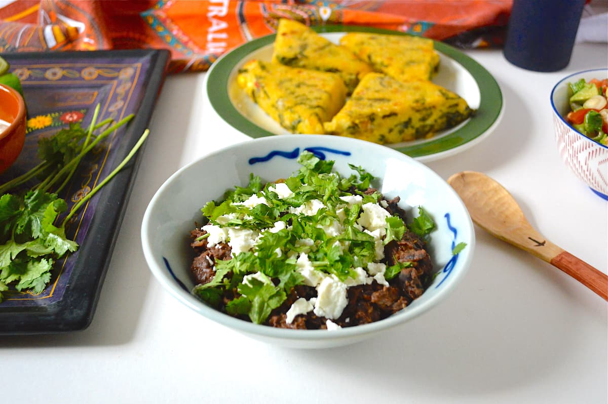 A bowl of Mexican refried beans topped with feta cheese and coriander. Other food items in the background.