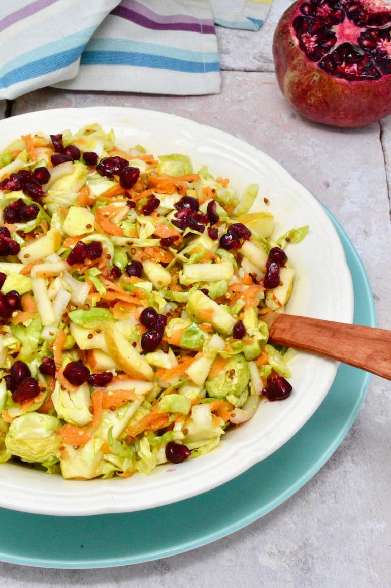 A bowl of Brussels sprout slaw with pomegranate seeds and a wooden spoon sitting on a blue plate.