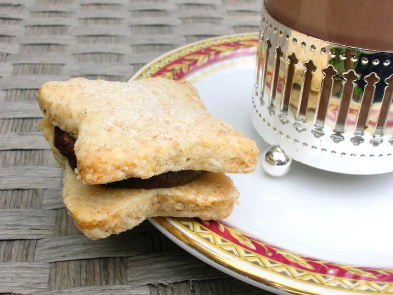 Vanilla biscuit sandwiched with chocolate ganache on the side of a plate containing a glass of hot chocolate.