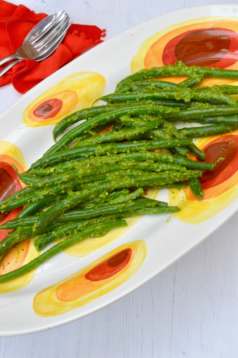 Pesto green beans served on a patterned oval platter with serving spoon and fork to the side.
