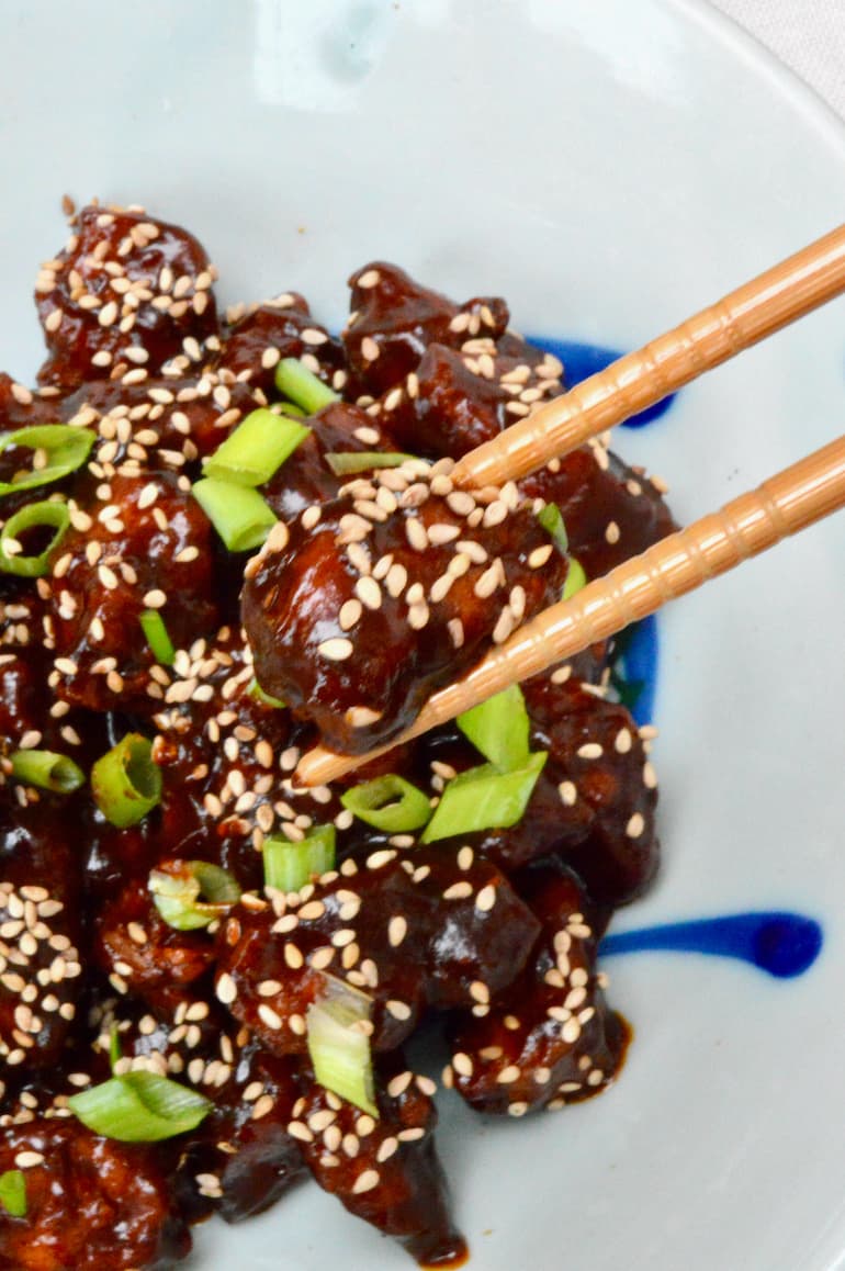 A piece of crispy air fryer tofu with sticky sesame sauce held by a pair of chopsticks.