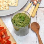 An open jar of Italian green sauce (bagnèt vert) with wooden spoon, knives and crackers.