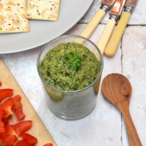 An open jar of Italian green sauce (bagnèt vert) with wooden spoon, knives and crackers.