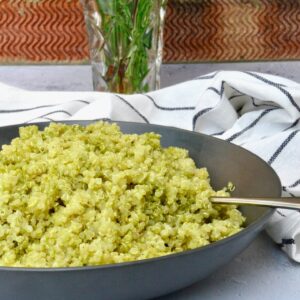 A grey bowl of fluffy quinoa with pesto. A striped grey cloth and a vase in the background.