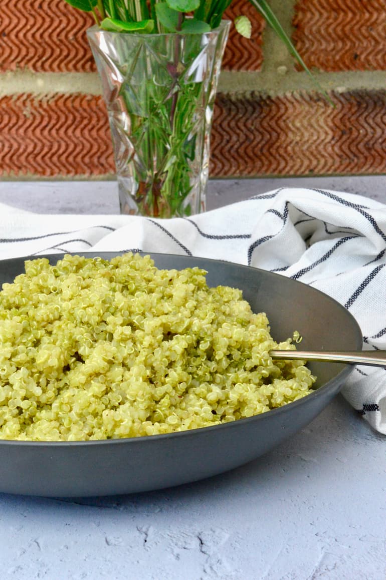 A grey bowl of fluffy quinoa with pesto. A striped grey cloth and a vase in the background.