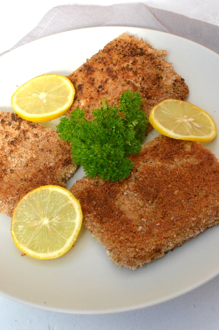3 pieces of vegan tofu schnitzel on a plate with slices of lemon and a sprig of parsley.
