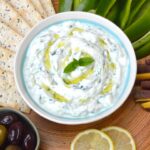A bowl of homemade tzatziki with crudités, crackers, olives and lemon slices.