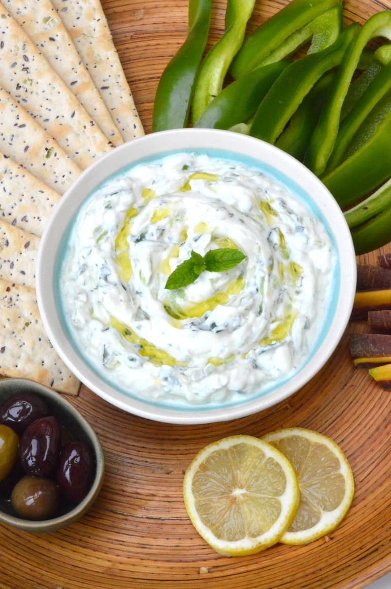 A bowl of homemade tzatziki with crudités, crackers, olives and lemon slices.