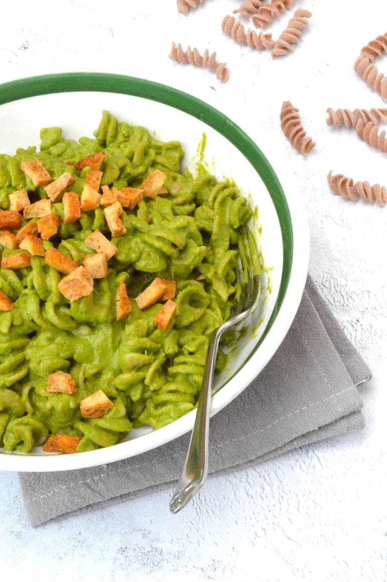 Partial bowl of pasta covered in green kale sauce with tofu and fork.