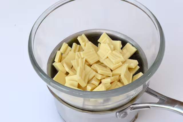 Broken white chocolate pieces in bowl over pan of hot water.