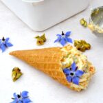 Tub of no-churn apricot ice cream with pistachio brittle and a scoop in waffle cone.