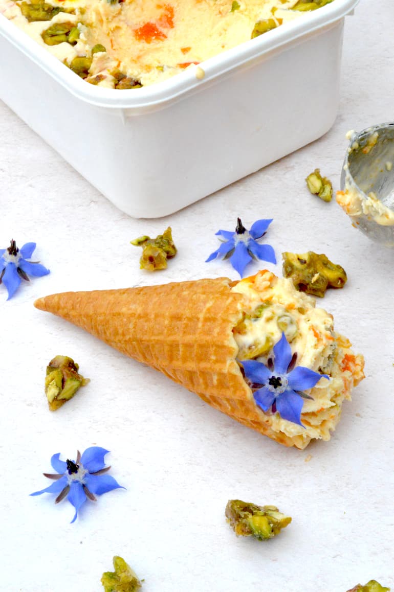 Tub of no-churn apricot ice cream with pistachio brittle and a scoop in waffle cone.