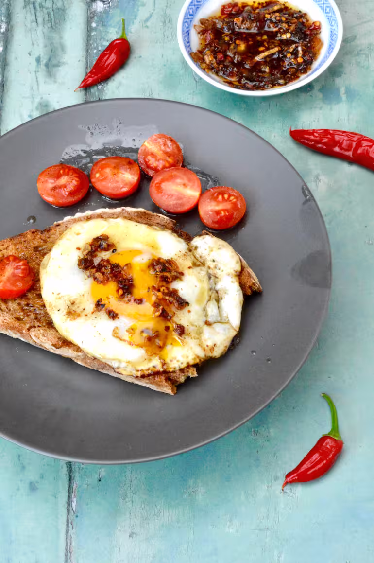 Homemade crispy chilli oil on top of fried egg on toast with a side of tomatoes.