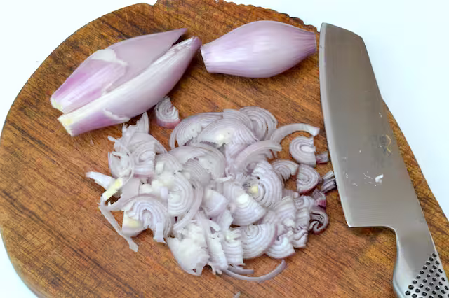 Peeled shallots on board with knife, one some finely sliced.