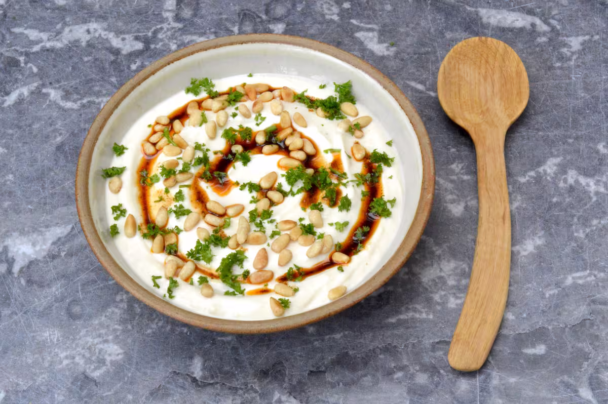 Bowl of tahini yoghurt sauce with spiced oil, toasted pine nuts and chopped parsley.