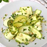 Ribbons of marinated courgettes curled on plate with herbs and toasted pine nuts.