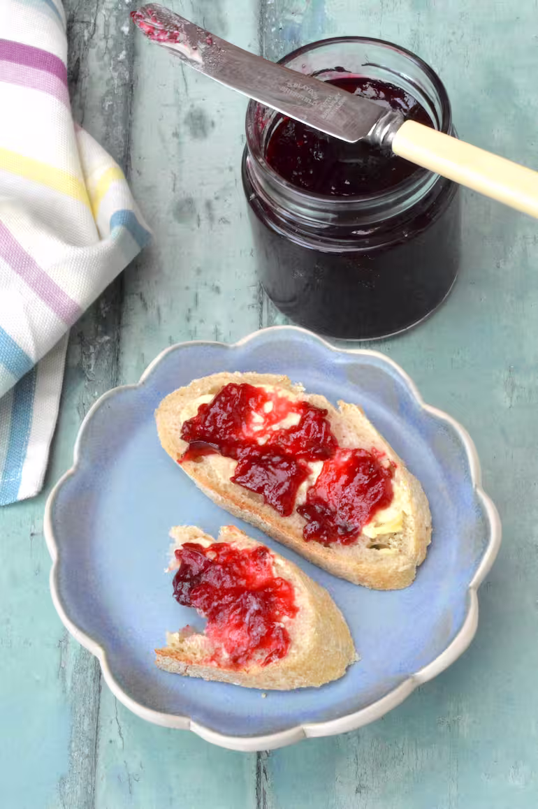 Damson jam spread on bread and butter with jam jar and knife.