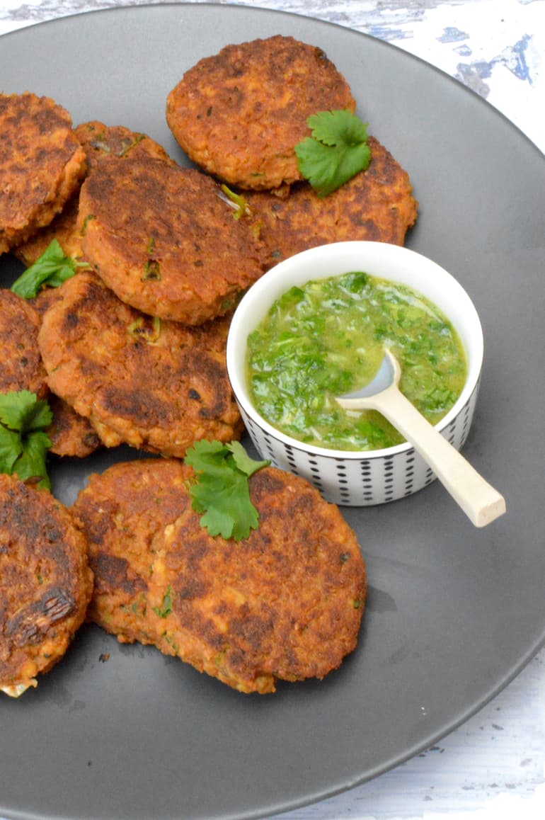 Vegan sweet potato tofu patties piled on plate with spoon inserted in bowl of green sauce.