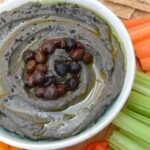 A bowl of black hummus topped with black chickpeas with crackers and crudités.